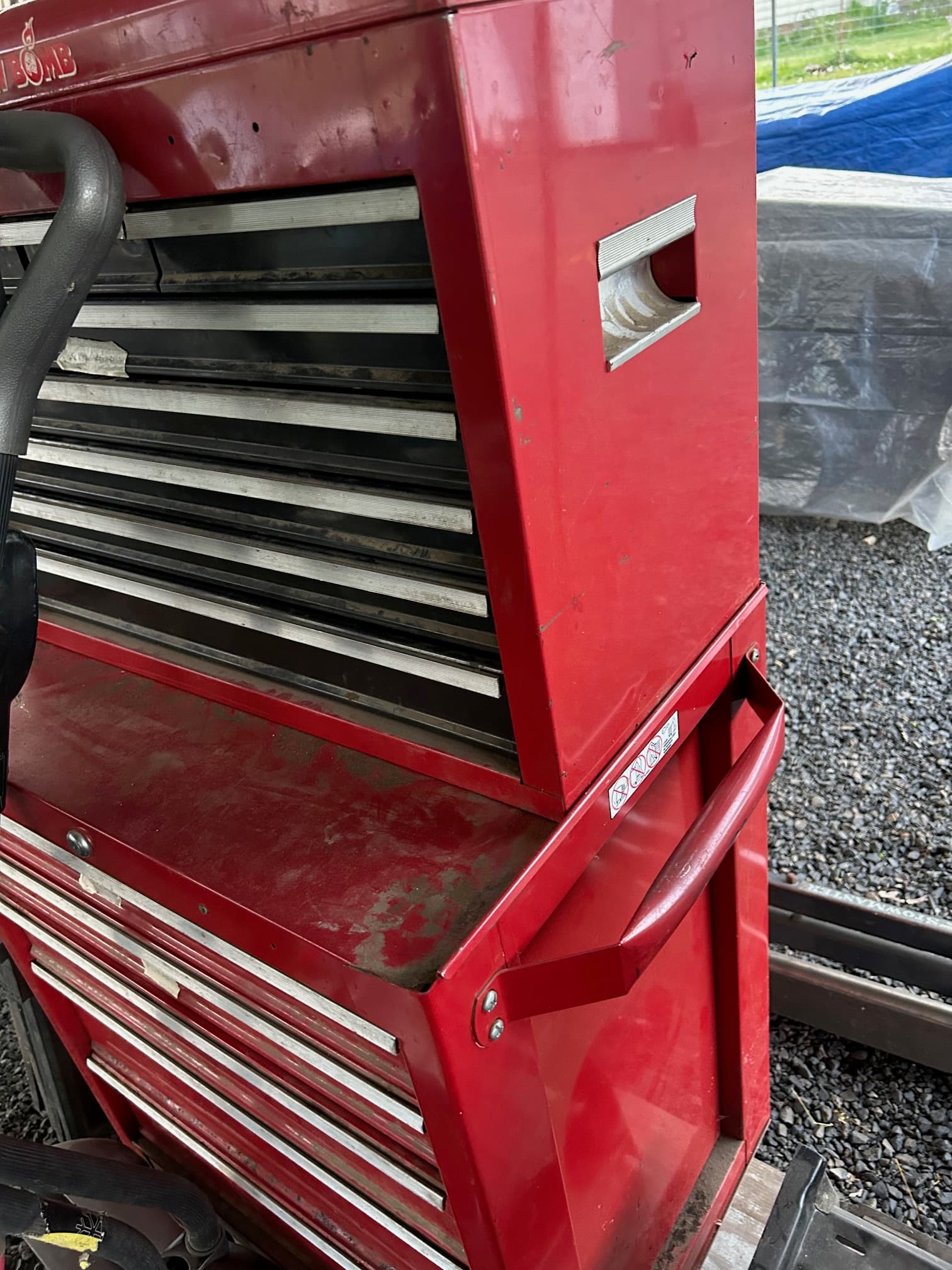 Red metal tool chest with multiple drawers and a top handle sitting on gravel.