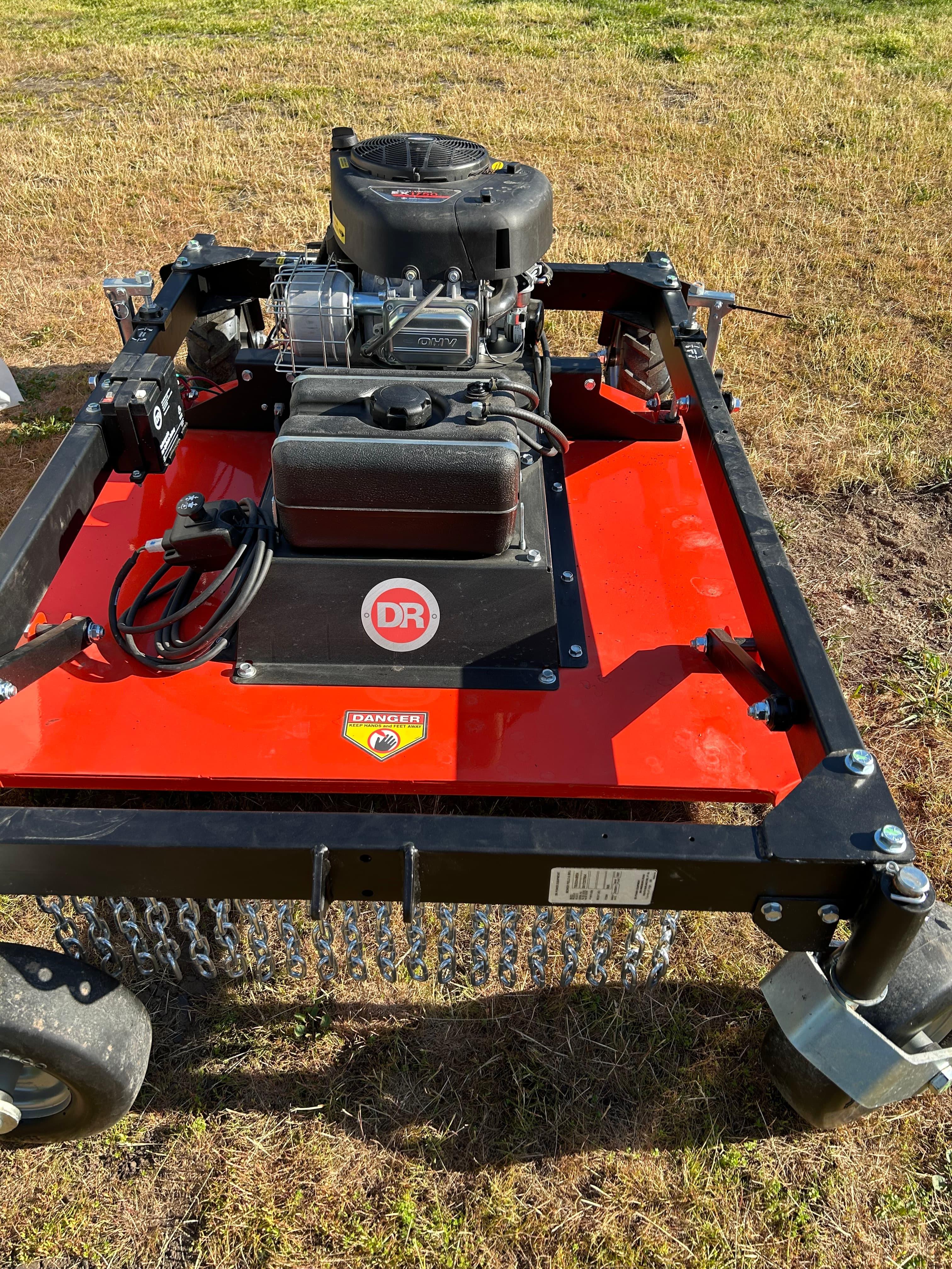 Red and black DR tow-behind field mower with safety chains sitting on dry grass.