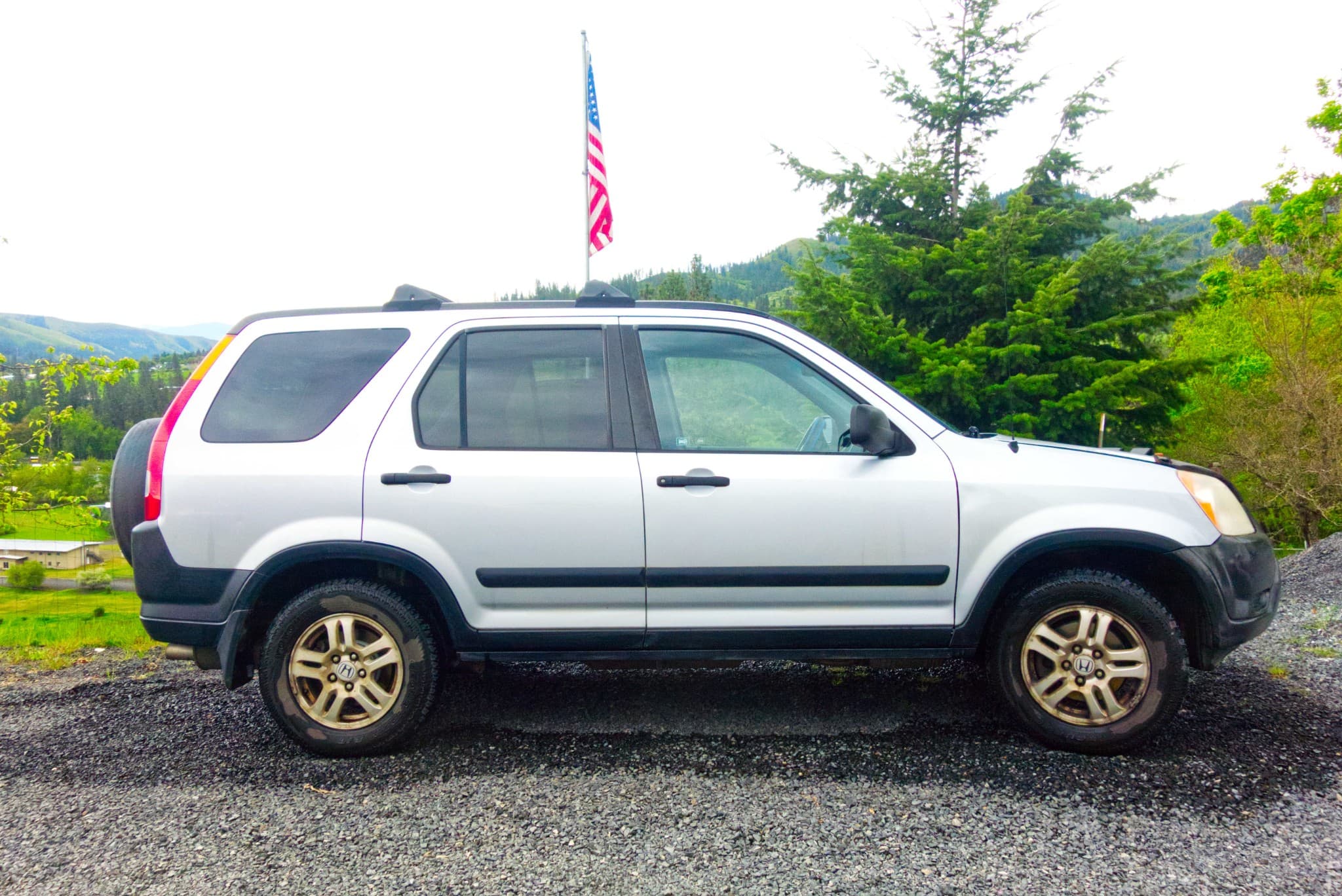 Silver Honda CR-V parked on gravel with an American flag and green hills behind it.