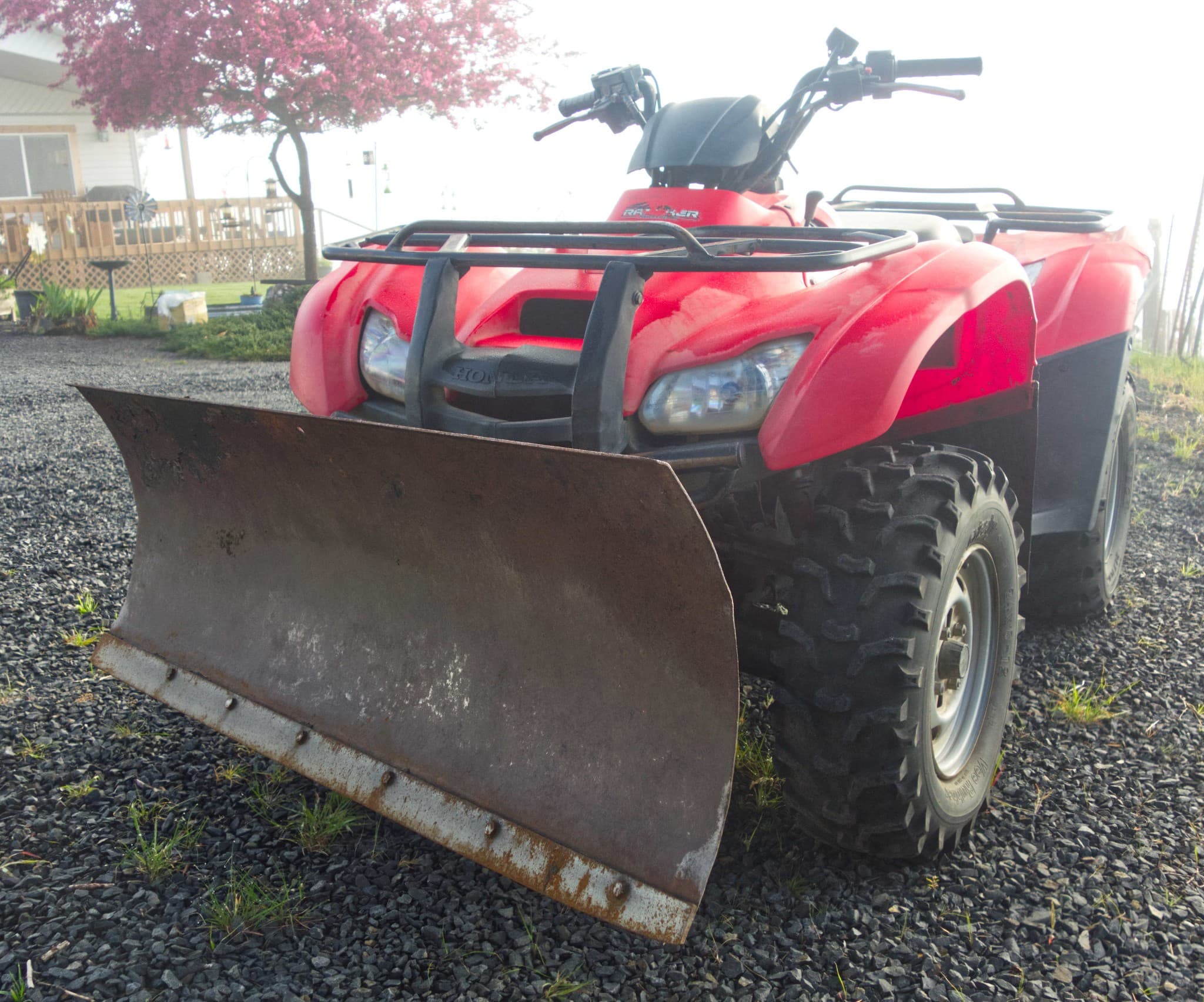 Red Honda ATV with a large rusty snow plow attachment parked on gravel.