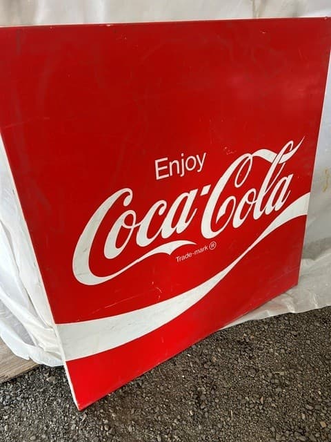 Square red sign featuring the white Coca-Cola logo, 'Enjoy' text, and a dynamic ribbon.