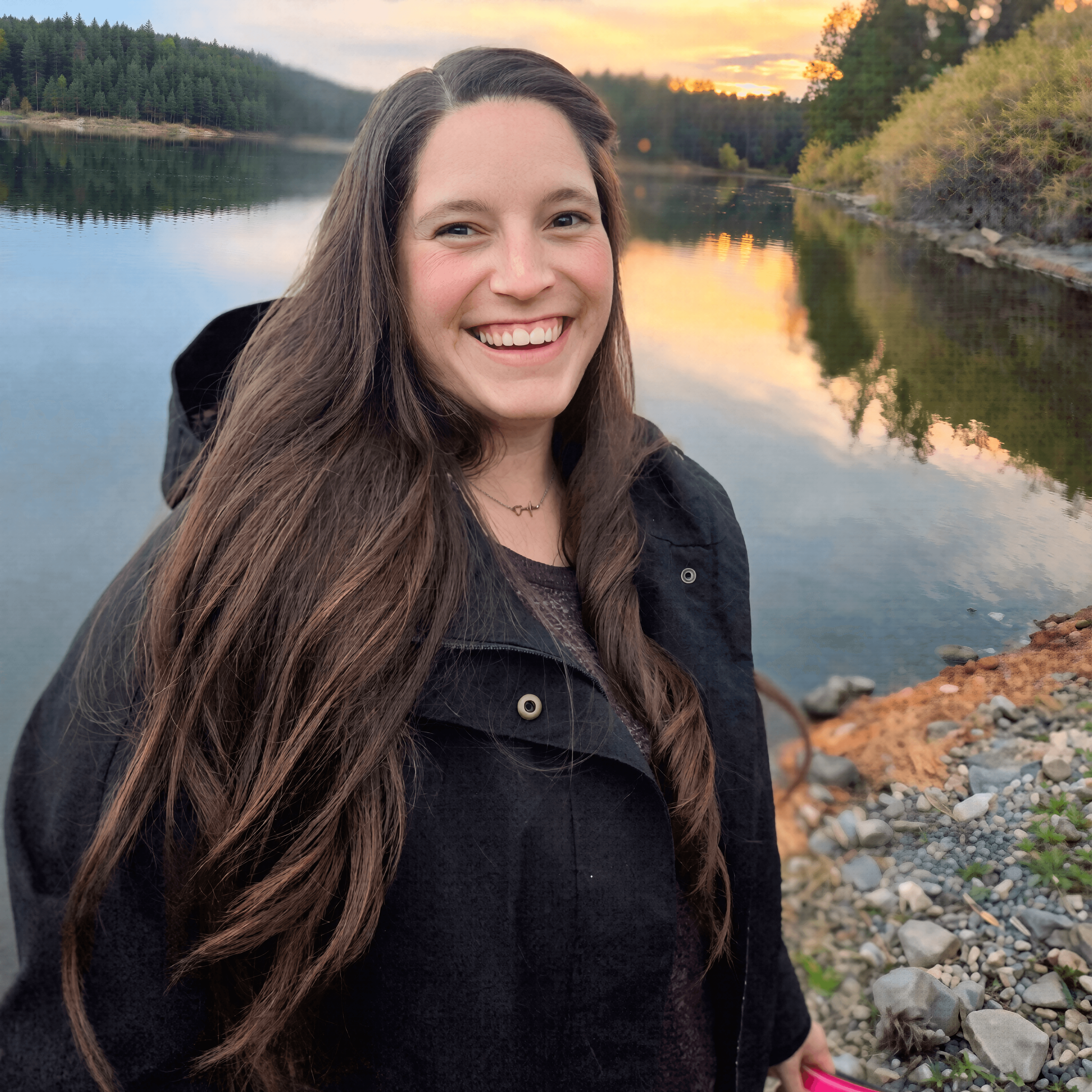 Smiling woman with long brown hair stands before a calm lake reflecting a sunset.