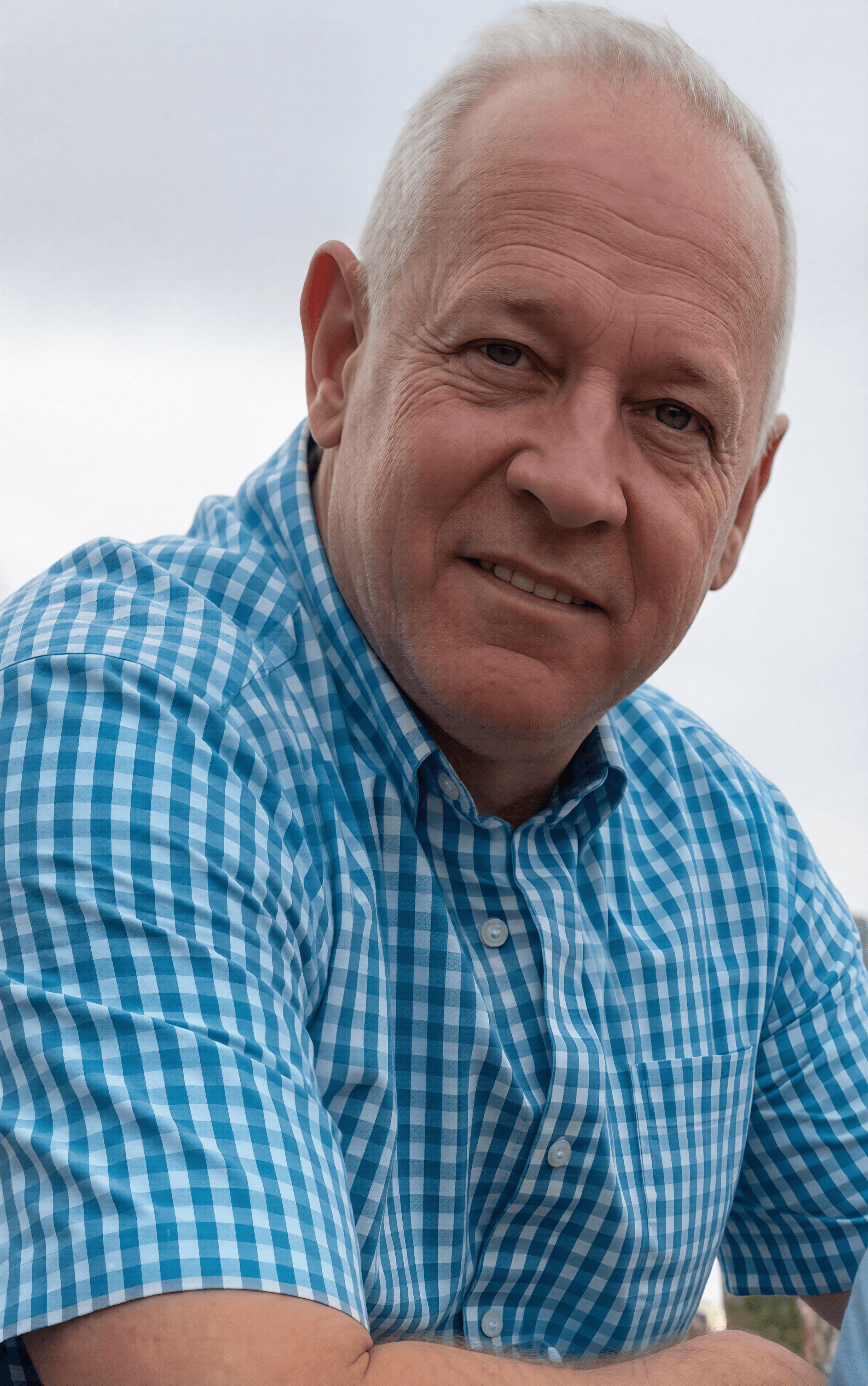 Smiling senior man with short white hair wearing a blue and white checkered shirt.