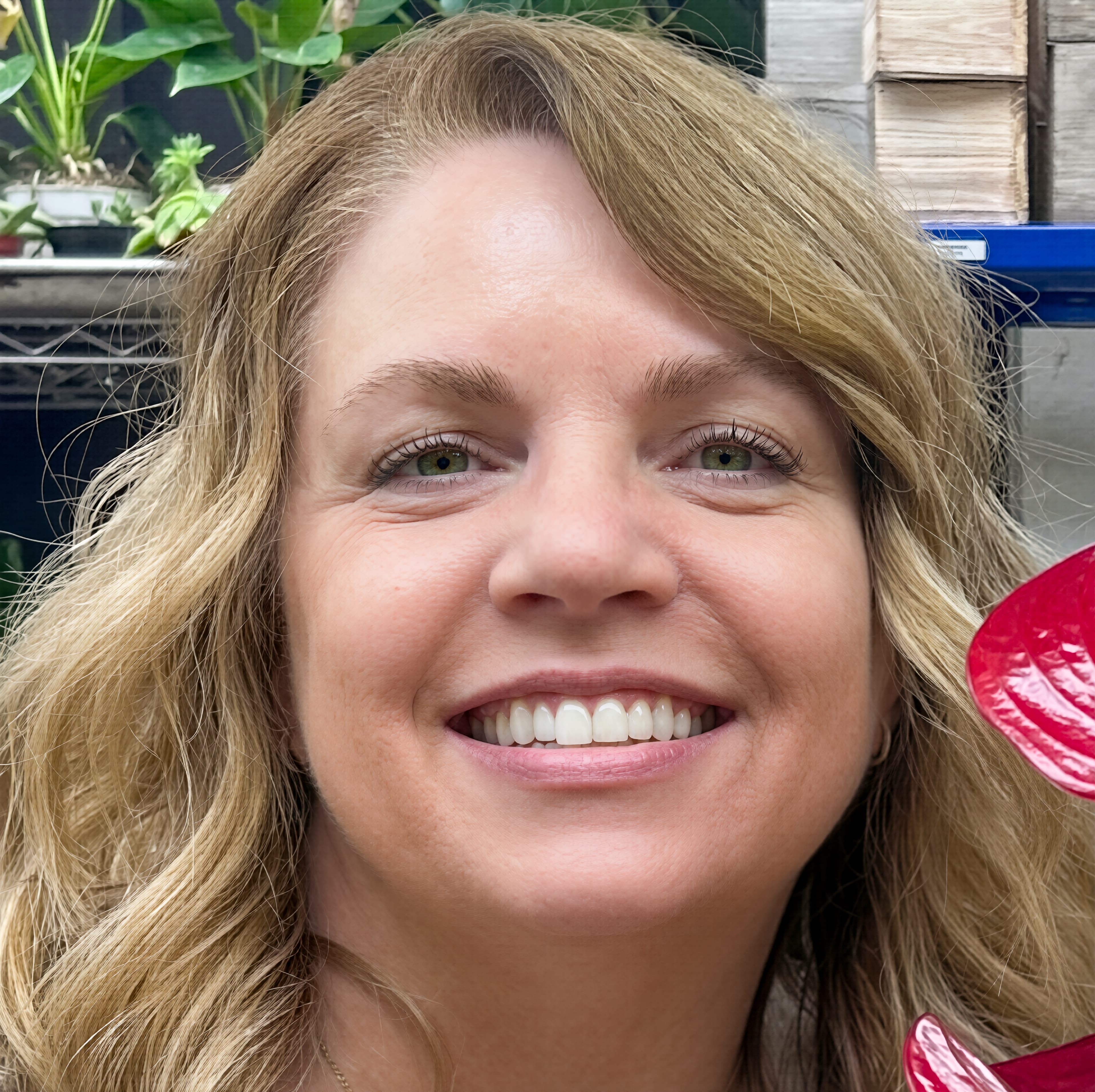 Close-up portrait of a smiling blonde woman with green eyes, surrounded by lush green plants.