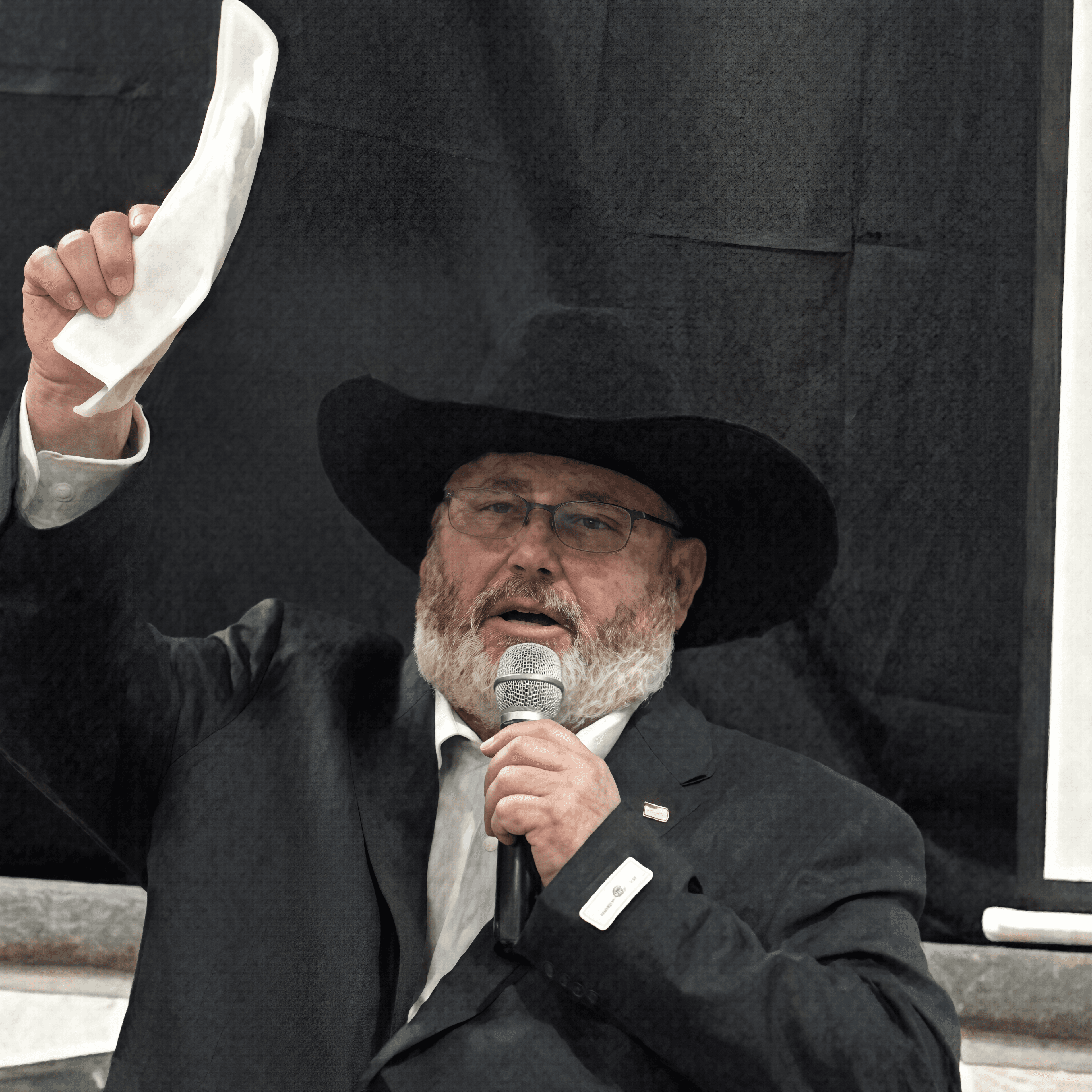 Bearded man in a black cowboy hat and suit speaking into a microphone, holding paper.