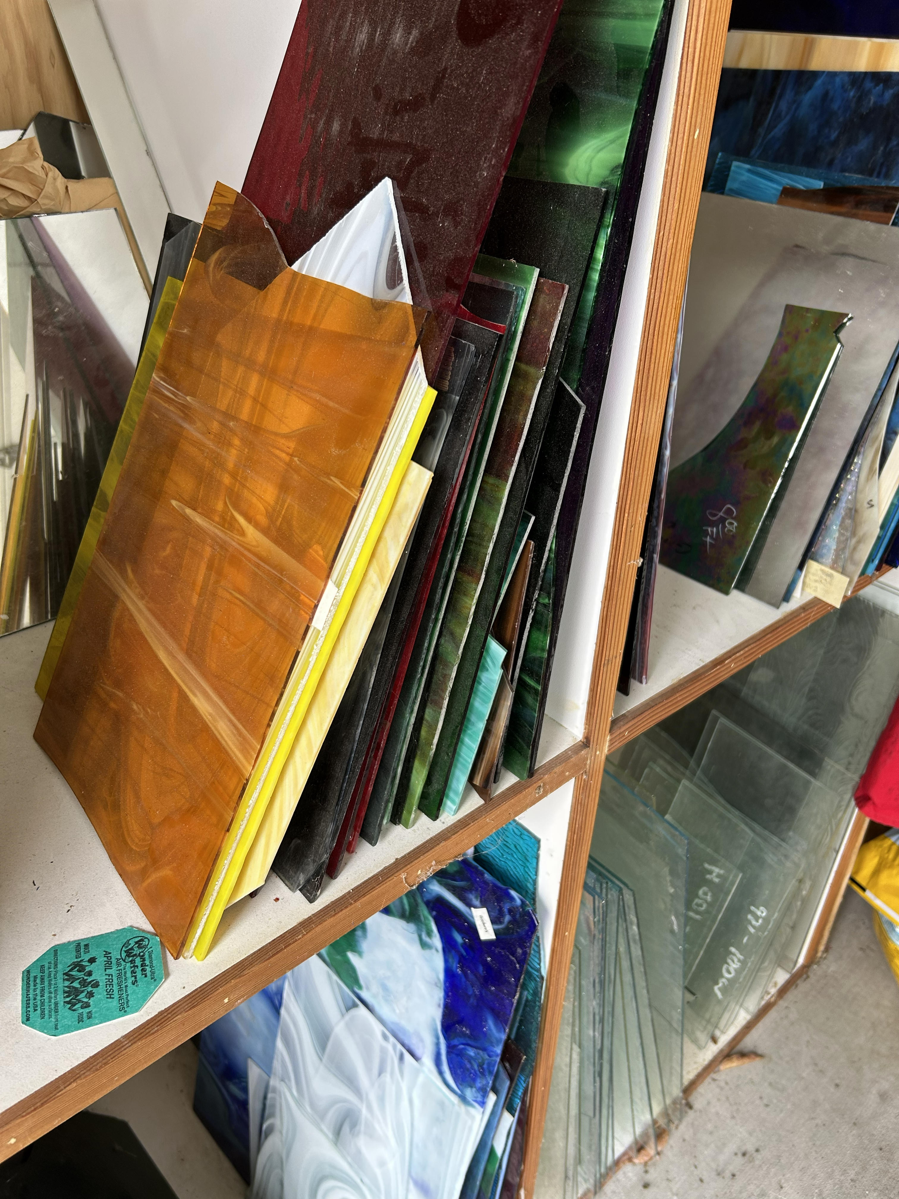Vibrant sheets of amber, green, and blue stained glass leaning against a wooden shelf.